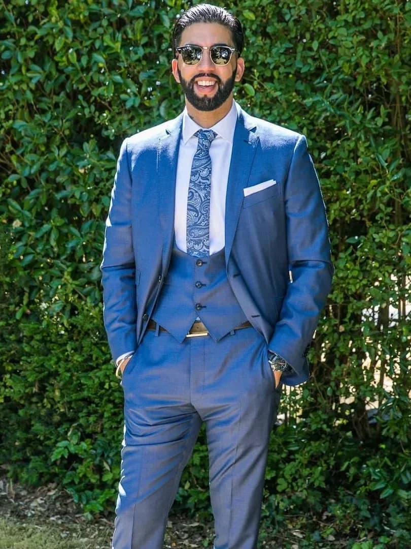 A man smiling outdoors in a blue three-piece suit with a patterned tie and white pocket square